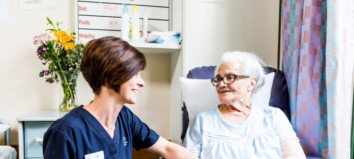 A Mercy Health female worker with short brown hair sits to the left of an elderly resident who holds her hand. They smile at each other.