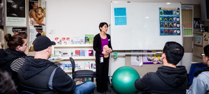 A long shot of a woman standing next to a green yoga ball in front of a room of people sitting and watching her.