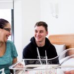 A man and woman sit looking at each other smiling in front of a sleeping baby in a neonatal bed.