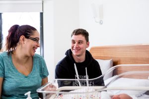A man and woman sit looking at each other smiling in front of a sleeping baby in a neonatal bed.