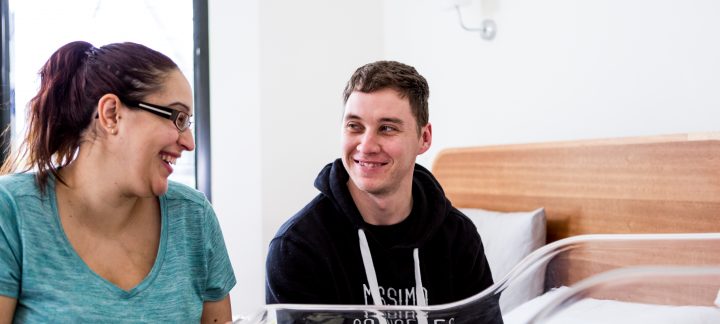 A man and woman sit looking at each other smiling in front of a sleeping baby in a neonatal bed.