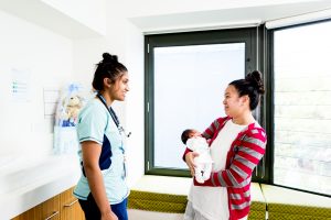 A female Mercy Health worker with a stethoscope smiles at a smiling mother holding a new born baby.