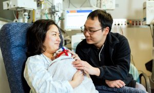 A woman sits holding a baby, smiling at a man sitting next to her.
