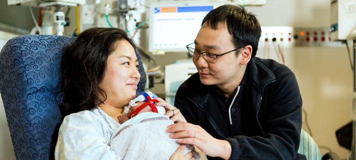 A woman sits holding a baby, smiling at a man sitting next to her.