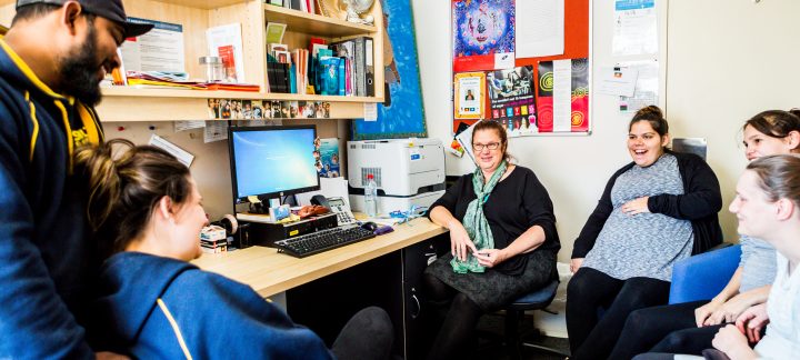 One man stands and five women sit and chat around a computer desk in a small office