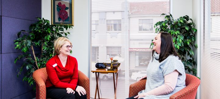 Two women sit in red arm chairs in a small room talking and laughing.