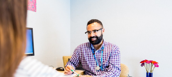 An over the shoulder photo of a man in a checked button up shirt wearing glasses sitting and smiling at a woman he is speaking to.