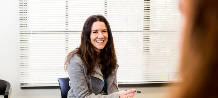 A young woman with long brown hair sits in a chair with a clipboard and pen, laughing.