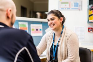 A woman wearing a blue lanyard smiles while she chats with her colleague as they sit behind a medical reception desk at the Ursula Fayne Centre.