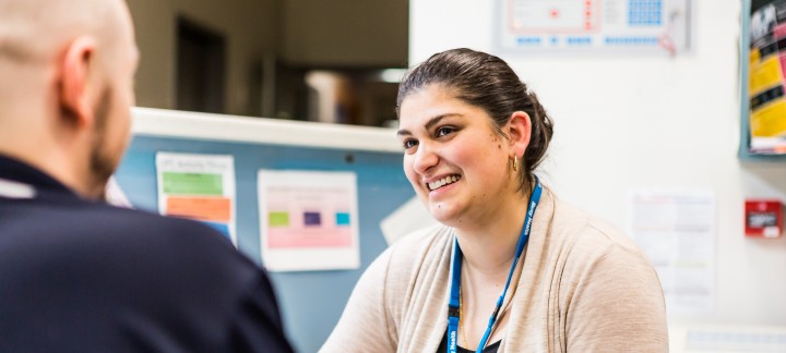 A woman wearing a blue lanyard smiles while she chats with her colleague as they sit behind a medical reception desk at the Ursula Fayne Centre.