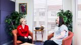 Two women sit in red arm chairs in a small room talking and laughing.