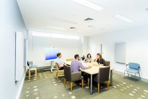 Two men and two women in casual corporate clothing sit at a board table in a white room having a meeting.