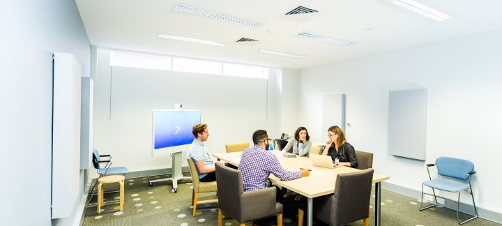 Two men and two women in casual corporate clothing sit at a board table in a white room having a meeting.