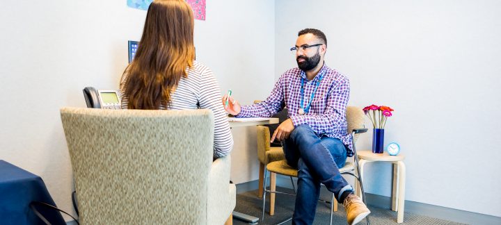 A long shot of a man in a checked button up shirt wearing glasses sitting with a woman at a desk.