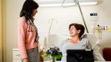 A female patient sits in bed in front of flowers and a propped up tablet, looking up at a female doctor in casual clothing, smiling.