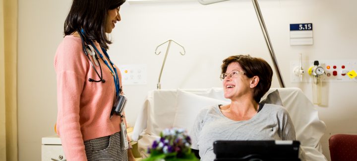 A female patient sits in bed in front of flowers and a propped up tablet, looking up at a female doctor in casual clothing, smiling.