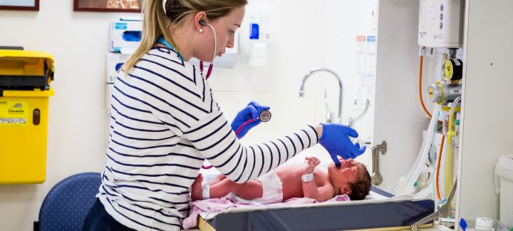 A medium long shot of a woman wearing blue gloves tending to a new born baby on a table.