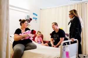 A long shot of a woman sitting on a hospital bed holding a baby while a woman stands and a man and a young child sit watching