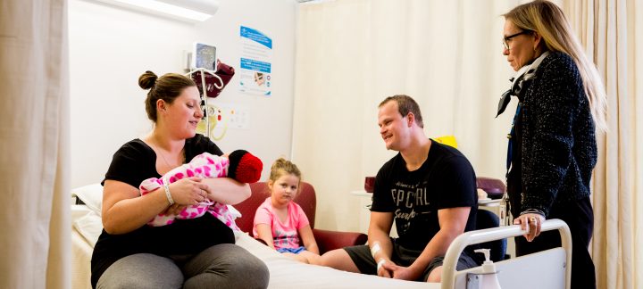 A long shot of a woman sitting on a hospital bed holding a baby while a woman stands and a man and a young child sit watching