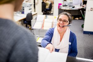 A woman sitting down wearing a headset is laughing and handing a booklet to the person standing in front of her