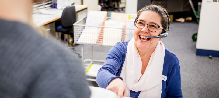 A woman sitting down wearing a headset is laughing and handing a booklet to the person standing in front of her