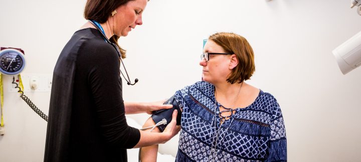 A female doctor stands next to a seated patient and takes her blood pressure