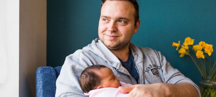 A man sits smiling out the window while holding a new born baby swaddled in pink
